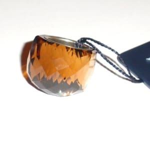 May include: A brown crystal ring with a silver band. The ring is on a white surface with a blue tag that says "SWAROVSKI" in white letters.