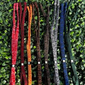 May include: A collection of seven knitted eyeglass straps in various colors, including red, orange, brown, gray, blue, and green. Each strap features a small white tooth-shaped charm. The straps are displayed against a green leafy background.