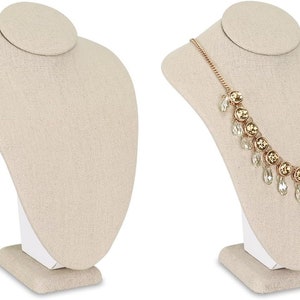 May include: Two beige fabric necklace display busts. One bust is empty, the other displays a gold-toned necklace with clear and gold-colored crystal accents. The busts have a curved shape and a small, round head support.