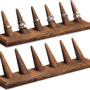May include: Two wooden ring displays with a dark brown finish. The displays have a rectangular base with six conical ring holders each. Several rings with various designs are displayed on the top ring holder.