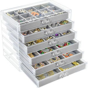 May include: A clear acrylic jewelry organizer with multiple drawers and compartments. The drawers are lined with gray velvet and filled with various pieces of jewelry, including rings, necklaces, and bracelets. The organizer has clear, crystal-like drawer pulls.