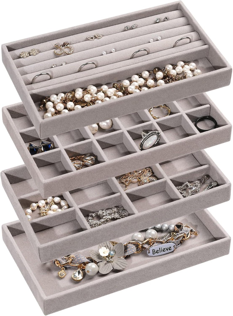 Op de afbeelding: Een set van vier stapelbare lichtgrijze fluwelen sieradenbakjes. De bovenste lade heeft lange sleuven voor ringen en oorbellen. De drie onderste lades zijn verdeeld in vierkante vakjes met diverse kettingen, armbanden, ringen en een bedel met het woord Believe.
