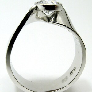 May include: A close-up of a white gold ring with a large, round, clear diamond. The ring has a modern, flowing design with a smooth, polished finish. The diamond is set in a bezel setting.