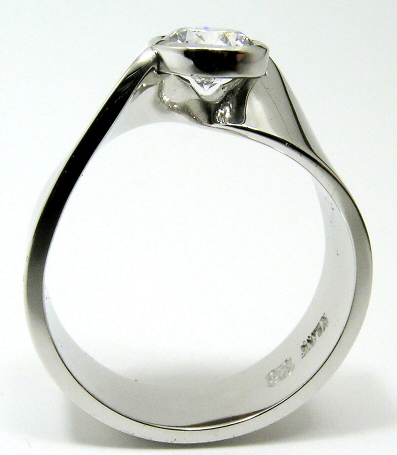 May include: A close-up of a white gold ring with a large, round, clear diamond. The ring has a modern, flowing design with a smooth, polished finish. The diamond is set in a bezel setting.