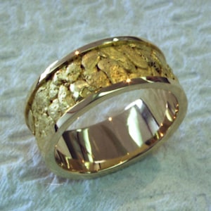 Może przedstawiać: Złoty pierścionek z teksturowanym paskiem z wzorem przypominającym samorodek. Pierścionek ma wypolerowane, gładkie wnętrze i jasną, odblaskową powierzchnię. Pasek ma szerokość około 1,3 cm, prezentując unikalną, organiczną estetykę.