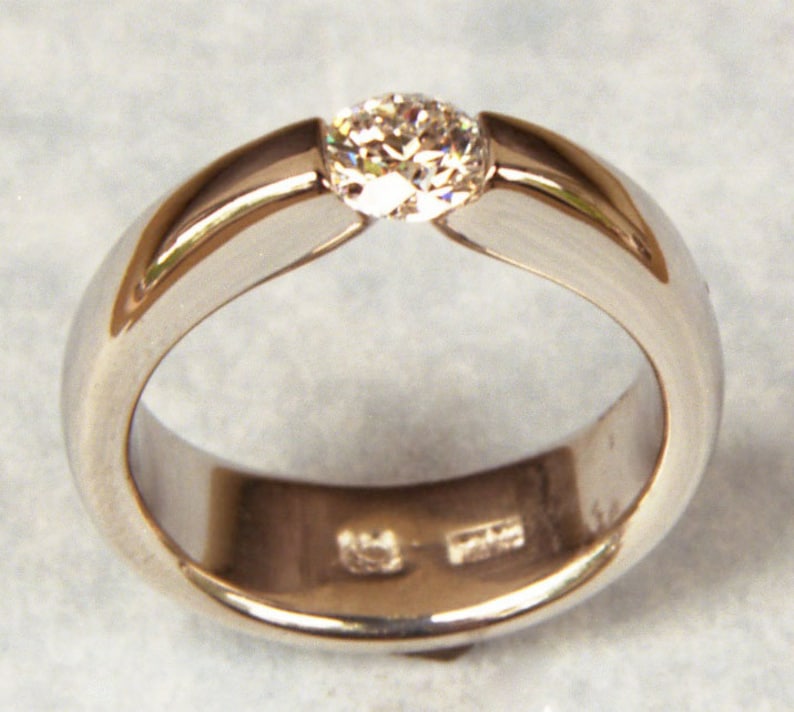 May include: A simple silver band ring with a single, round, clear diamond set in the centre.