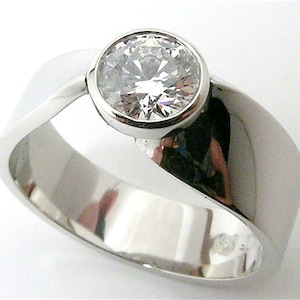 May include: A polished, silver-toned ring with a large, round, clear gemstone set in a bezel. The band has a smooth, curved design, creating a modern and elegant look. The gemstone sparkles, catching the light. A piece of jewelry.