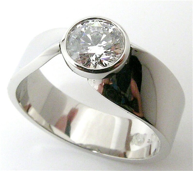 May include: A polished, silver-toned ring with a large, round, clear gemstone set in a bezel. The band has a smooth, curved design, creating a modern and elegant look. The gemstone sparkles, catching the light. A piece of jewelry.