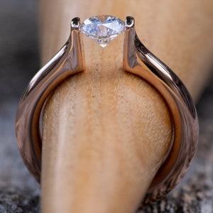May include: A rose gold engagement ring with a single, clear, round diamond set in a bezel setting.