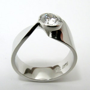 May include: A modern, silver-toned ring with a large, clear, round gemstone. The ring has a unique, flowing design, with a band that curves around the finger and supports the gemstone. The ring is set against a white background.