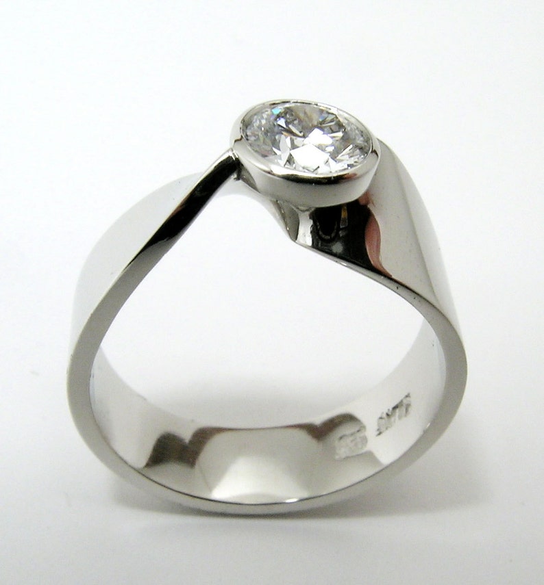 May include: A modern, silver-toned ring with a large, clear, round gemstone. The ring has a unique, flowing design, with a band that curves around the finger and supports the gemstone. The ring is set against a white background.
