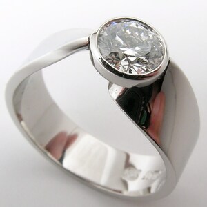 May include: A polished, silver-toned ring with a large, round, clear gemstone. The ring band has a flowing, asymmetrical design, with a smooth, reflective surface. The gemstone is set in a bezel setting, enhancing its brilliance. A piece of jewelry.