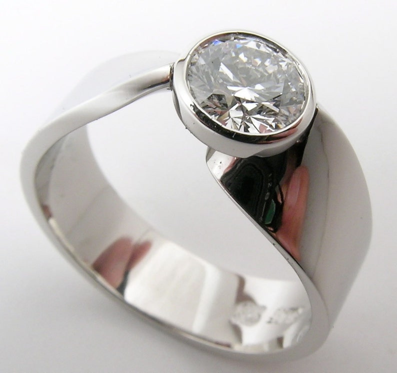 May include: A polished, silver-toned ring with a large, round, clear gemstone. The ring band has a flowing, asymmetrical design, with a smooth, reflective surface. The gemstone is set in a bezel setting, enhancing its brilliance. A piece of jewelry.