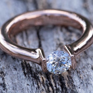 May include: A rose gold engagement ring with a single round diamond set in a unique prong setting.