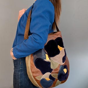 Puede incluir: Un bolso de hombro de cuero marrón con un diseño floral. El bolso presenta una mezcla de texturas y colores, incluyendo azul oscuro, beige y negro. La correa es de color marrón claro.