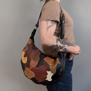 Peut inclure: Sac à bandoulière en cuir noir avec un motif patchwork dans les tons marron, beige et crème. Le sac a une forme incurvée et une longue bandoulière réglable avec des détails dorés. Le sac est porté à l'épaule.