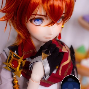May include: A close-up of a doll's face with blue eyes and red hair. The doll is wearing a black glove and a red and white scarf. The doll's eyes are closed and the doll is looking down. The doll is wearing a blue contact lens with the text "shinesworkshop" printed on it.