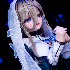 May include: A detailed doll with a white veil adorned with floral accents. The doll has blonde hair, blue eyes, and a tear-like mark on its cheek. It wears a white and teal dress with gold accents.