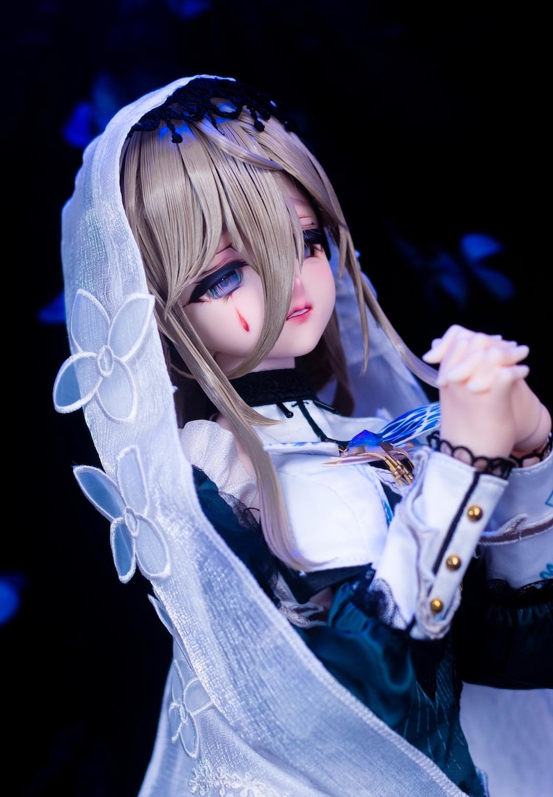 May include: A detailed doll with a white veil adorned with floral accents. The doll has blonde hair, blue eyes, and a tear-like mark on its cheek. It wears a white and teal dress with gold accents.