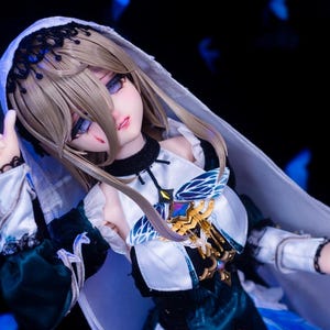 May include: A detailed doll with a pale complexion, wearing a white and teal dress with gold accents. The doll has long, light brown hair and is wearing a white veil. The doll's eyes are a striking blue, and it has a small wound on its face.