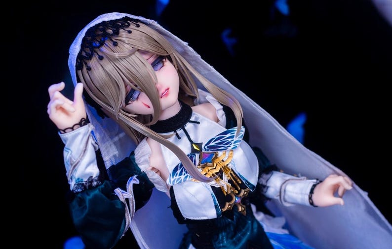 May include: A detailed doll with a pale complexion, wearing a white and teal dress with gold accents. The doll has long, light brown hair and is wearing a white veil. The doll's eyes are a striking blue, and it has a small wound on its face.
