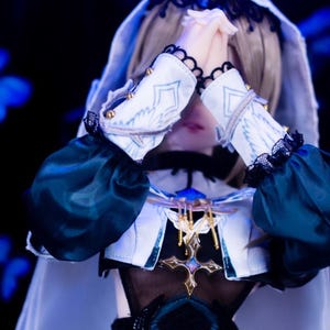 May include: A doll dressed in a white and teal outfit with a hood and sleeves. The doll's hands are clasped in prayer. The outfit has gold accents and black lace trim. The word "Pray" is at the bottom of the image.