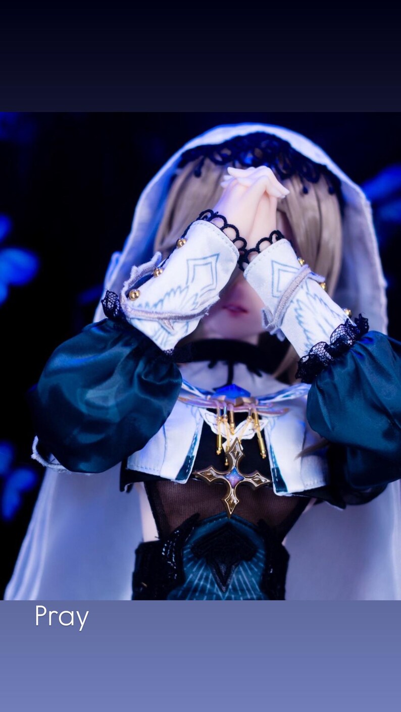 May include: A doll dressed in a white and teal outfit with a hood and sleeves. The doll's hands are clasped in prayer. The outfit has gold accents and black lace trim. The word "Pray" is at the bottom of the image.