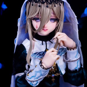 May include: A detailed doll with a pale complexion, wearing a white hooded veil with black lace trim. The doll has long, light brown hair and is dressed in a white and teal outfit with gold accents. A tear is visible on the doll's cheek.
