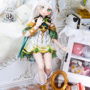 May include: A white haired doll with green eyes wearing a white and green dress with gold accents and white boots with gold accents. The doll is standing on a white surface.