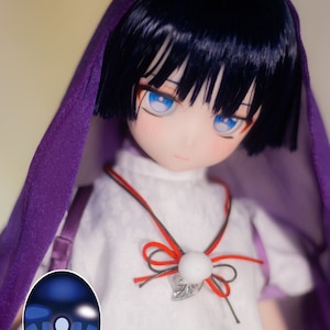 May include: A doll with blue eyes and black hair wearing a white shirt with a red and black bow and a purple hooded cloak. The doll is wearing blue eye contacts with a white rim.