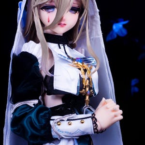 May include: A detailed doll with a pale complexion, long blonde hair, and a tear-shaped mark under one eye. The doll wears a white and dark green outfit with gold accents and a white veil. The doll's hands are clasped, and the background is dark.