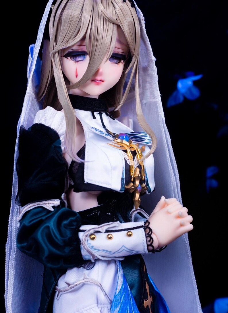 May include: A detailed doll with a pale complexion, long blonde hair, and a tear-shaped mark under one eye. The doll wears a white and dark green outfit with gold accents and a white veil. The doll's hands are clasped, and the background is dark.