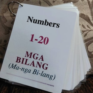 Może przedstawiać: Zestaw fiszek z tytułem "Numbers 1-20" w czcionce czarnej i czerwonej. Poniżej tekst "MGA BILANG (Ma-nga Bi-lang)" jest wydrukowany mniejszą, ciemną czcionką. Karty są spięte srebrnym metalowym kółkiem.