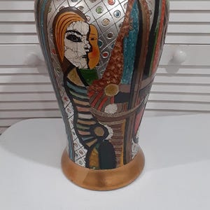 May include: A decorative vase with a metallic gold rim and base. The vase features a painted design with figures and geometric shapes in silver, brown, and blue. The artwork depicts two faces and other abstract elements.