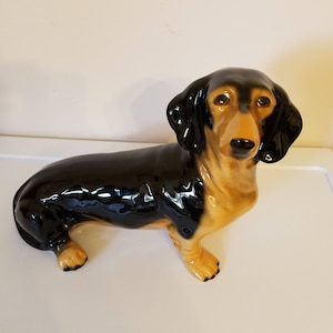 May include: A black and tan ceramic dachshund figurine. The figurine is sitting with its head turned to the side and its ears perked up.