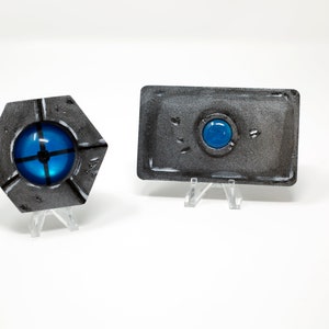 May include: Two small, gray, metal, decorative objects with blue glass accents. The object on the left is hexagonal shaped and the object on the right is rectangular.