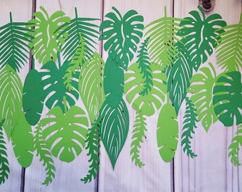 May include: A collection of green paper leaves, cut in various shapes, including palm leaves, ferns, and other tropical foliage. The leaves are arranged in a row on a white wooden background.