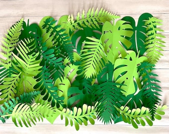 May include: A collection of green paper leaves, including ferns, palm leaves, and monstera leaves, arranged on a light wood background.