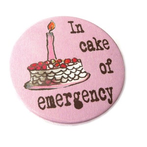 Op de afbeelding: Een ronde, roze button met de tekst "In cake of emergency" in bruine letters. De button heeft een handgetekende illustratie van een taart met een brandende kaars.