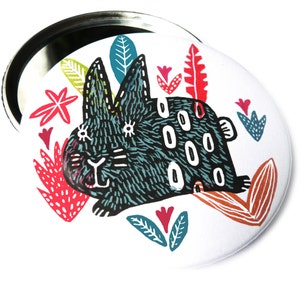 May include: A white round compact mirror with a black rabbit illustration surrounded by colorful flowers and leaves. The rabbit has white eyes and a black nose.