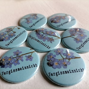 Op de afbeelding: Zeven ronde blauwe en witte knopen met de Duitse zin "Vergissmeinnicht" (vergeet-me-nietje) in zwart gedrukt op elke knop. Elke knop heeft een geperst bloemenmotief van vergeet-me-nietjes.