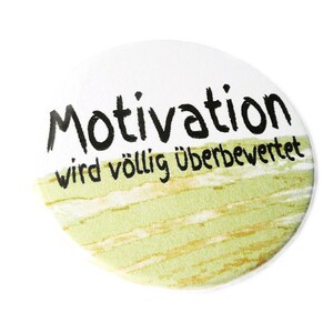 Könnte beinhalten: Ein weißer runder Knopf mit einem grün-weiß gestreiften Hintergrund. Der Text "Motivation wird völlig überbewertet" ist in Schwarz auf dem Knopf gedruckt.