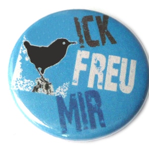 Könnte beinhalten: Ein blauer Knopf mit einer schwarzen Vogelsilhouette und dem Text "ICK FREU MIR" in weißer und schwarzer Schrift.