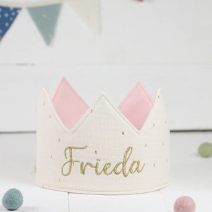 Könnte beinhalten: Eine cremefarbene Stoffkrone mit dem Namen "Frieda" in goldener Schrift gestickt. Die Krone hat ein rosa Innenfutter und ist mit kleinen goldenen Punkten verziert. Der Hintergrund zeigt eine weiße Holzoberfläche und bunte Wimpel.