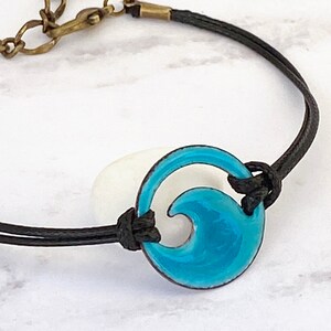 Puede incluir: Un brazalete de cordón negro con un dije de onda de esmalte azul turquesa. El dije es redondo y tiene un diseño de onda recortada.