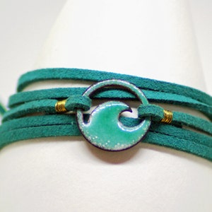 Puede incluir: Un brazalete de envoltura de gamuza azul turquesa con un acento de metal dorado y un dije de onda de esmalte azul turquesa.