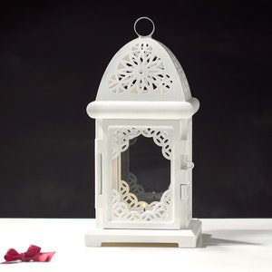 May include: White metal lantern with intricate cut-out designs and a glass panel. The lantern has a domed top and a square base.