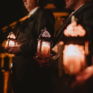 Puede incluir: Dos personas con traje sostienen linternas de metal adornadas con velas encendidas en su interior. Las linternas tienen diseños intrincados y son de un color dorado cálido.