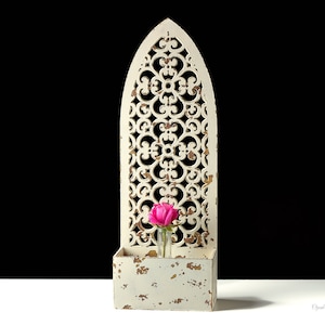 Peut inclure: Une décoration murale en bois blanc avec un motif découpé décoratif et une petite étagère. Une seule rose rose est dans un vase en verre sur l'étagère.