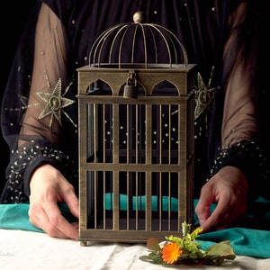 May include: A gold metal birdcage with a padlock on the door. The cage is sitting on a white surface with a green cloth behind it. A small bouquet of orange flowers is in front of the cage.